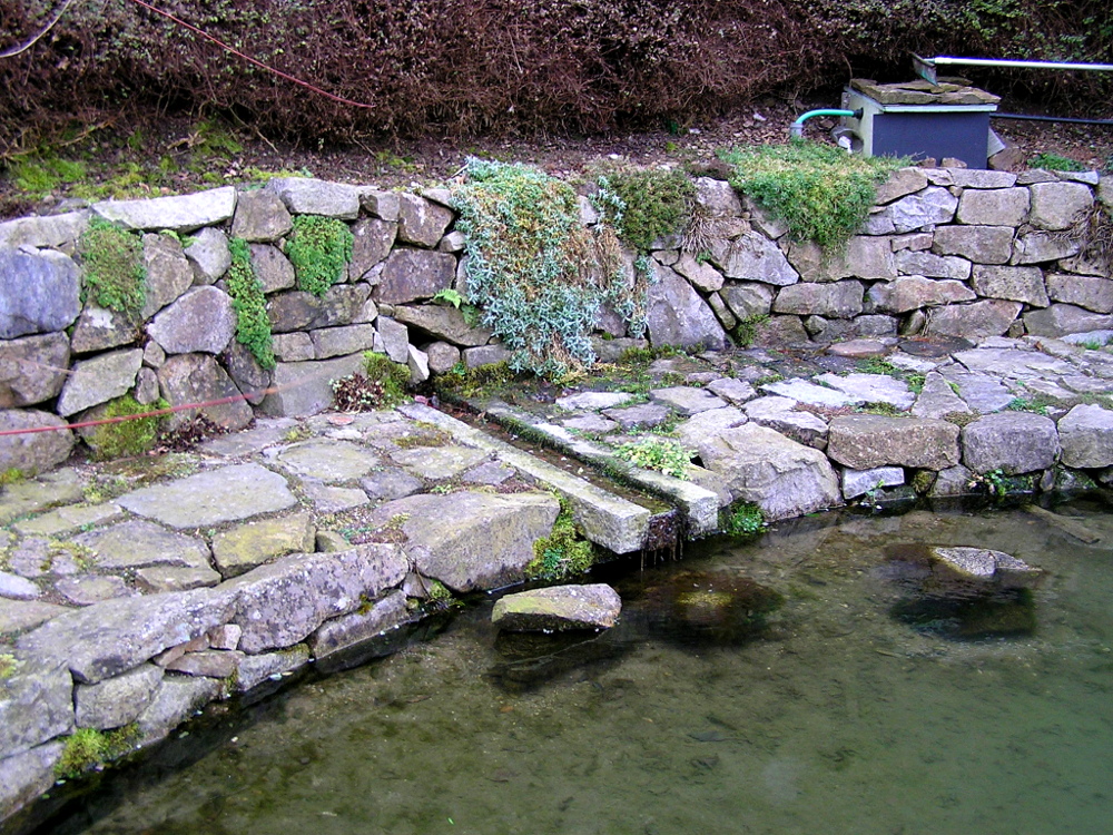 Einfassung einer natürlichen Quelle mit einer Granitrinne in den natürlichen Gartenteich und umlaufende Granit-Findling-Mauer und -Pflaster